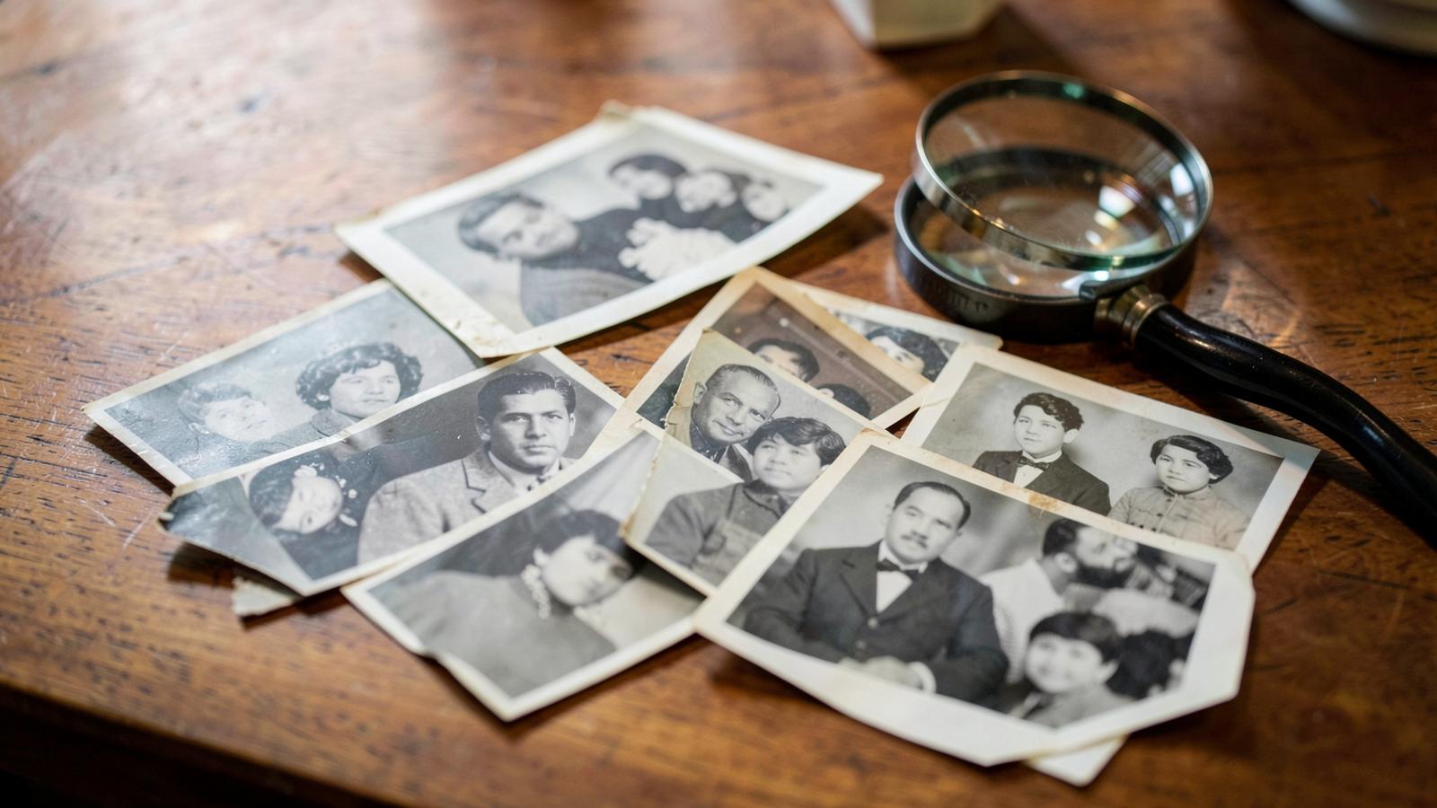 Several old black and white portraits spread on a wooden table next to a magnifying glass